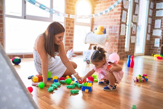 Young beautiful teacher and toddler sitting on the floor playing with building blocks toy at kindergarten