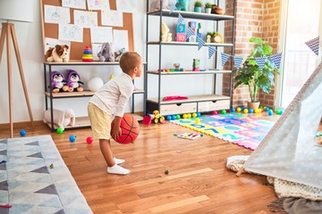 Beautiful blonde toddler holding basketball ball around lots of toys at kindergarten