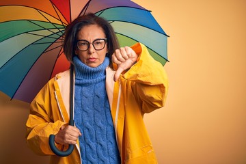 Middle age woman wearing yellow raincoat under colorful umbrella over isolated background with...