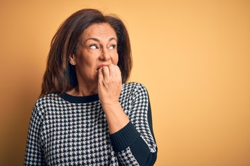 Middle age beautiful woman wearing casual sweater over isolated yellow background looking stressed and nervous with hands on mouth biting nails. Anxiety problem.