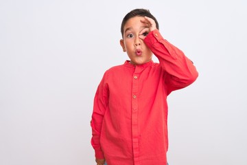 Beautiful kid boy wearing elegant red shirt standing over isolated white background doing ok gesture shocked with surprised face, eye looking through fingers. Unbelieving expression.