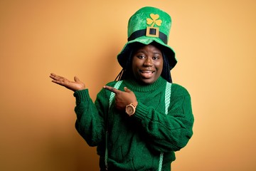 Plus size african american woman with braids wearing green hat with clover on st patricks day amazed and smiling to the camera while presenting with hand and pointing with finger.
