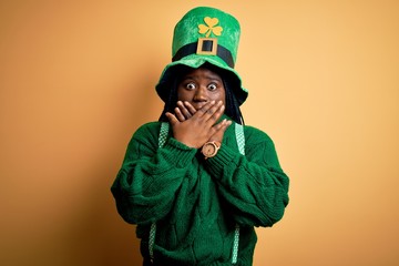 Plus size african american woman with braids wearing green hat with clover on st patricks day shocked covering mouth with hands for mistake. Secret concept.