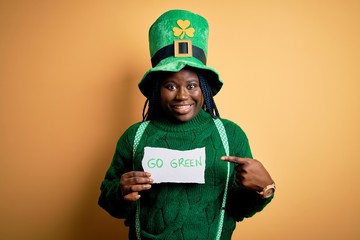 Plus size african american woman wearing green hat holding paper on saint patricks day very happy pointing with hand and finger