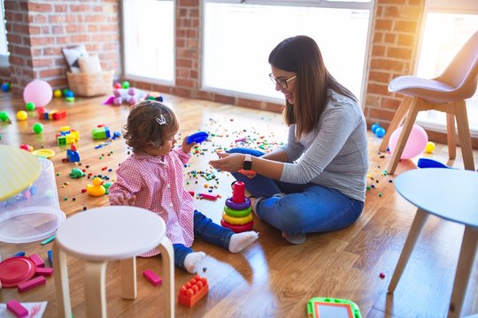 Young beautiful teacher and toddler sitting on the floor building pyramid using hoops at kindergarten