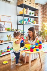 Young beautiful teacher and toddler playing with dishes, cutlery and cups toy on the table at...