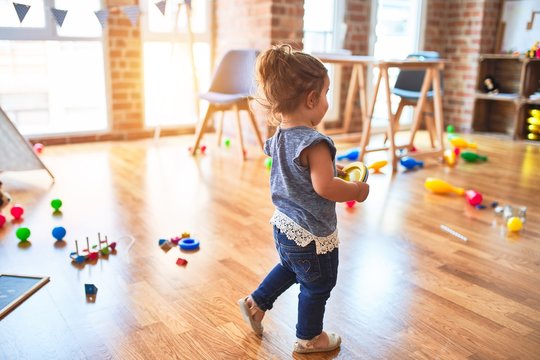 Beautiful toddler holding plastic dishes standing around lots of toys smiling at kindergarten