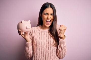 Young beautiful brunette woman holding piggy bank saving money for retirement annoyed and frustrated shouting with anger, crazy and yelling with raised hand, anger concept