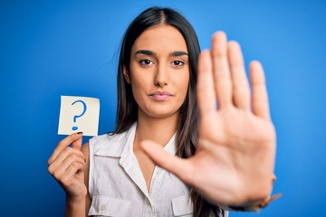 Young beautiful brunette woman holding paper with question mark symbol message with open hand doing stop sign with serious and confident expression, defense gesture