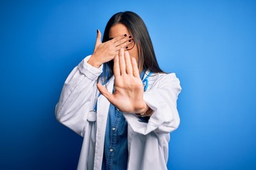 Young beautiful doctor woman wearing stethoscope and glasses over blue background covering eyes with hands and doing stop gesture with sad and fear expression. Embarrassed and negative concept.