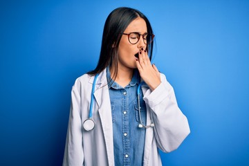 Young beautiful doctor woman wearing stethoscope and glasses over blue background bored yawning tired covering mouth with hand. Restless and sleepiness.