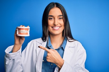 Young beautiful brunette dentist woman wearing coat holding plastic teeth prosthesis very happy pointing with hand and finger
