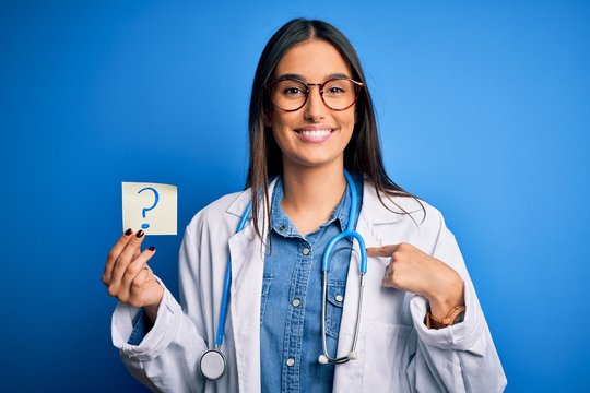 Young beautiful brunette doctor woman holding paper with question mark symbol message with surprise face pointing finger to himself