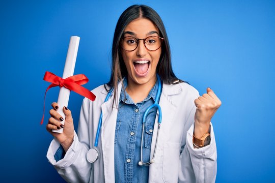 Young beautiful brunette doctor woman wearing glasses and coat holding diploma degree screaming proud and celebrating victory and success very excited, cheering emotion
