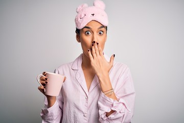 Young beautiful brunette woman wearing pajama and sleep mask drinking cup of coffee cover mouth...