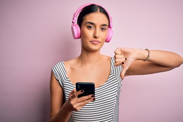 Naklejka premium Young beautiful brunette woman listening to music using headphones and smartphone with angry face, negative sign showing dislike with thumbs down, rejection concept