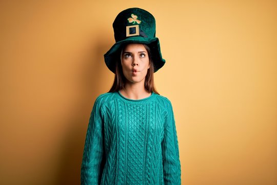 Young beautiful brunette woman wearing green hat with clover celebrating saint patricks day making fish face with lips, crazy and comical gesture. Funny expression.