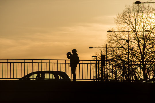 Mulhouse - France - 8 February 2020 - Silhouette of woman taking a photography on bridge with smartphone in hands by sunset