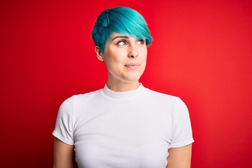 Young beautiful woman with blue fashion hair wearing casual t-shirt over red background smiling looking to the side and staring away thinking.
