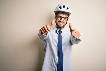 Young businessman wearing glasses and bike helmet standing over isolated white bakground approving doing positive gesture with hand, thumbs up smiling and happy for success. Winner gesture.