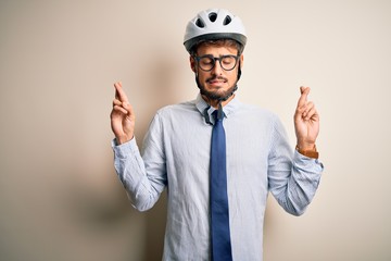 Young businessman wearing glasses and bike helmet standing over isolated white bakground gesturing finger crossed smiling with hope and eyes closed. Luck and superstitious concept.