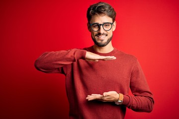 Young handsome man with beard wearing glasses and sweater standing over red background gesturing with hands showing big and large size sign, measure symbol. Smiling looking at the camera. Measuring