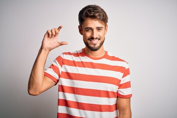 Fototapeta premium Young handsome man with beard wearing striped t-shirt standing over white background smiling and confident gesturing with hand doing small size sign with fingers looking and the camera. Measure