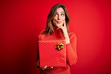 Young beautiful brunette woman holding birthday gift over isolated red background serious face...