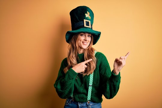Beautiful brunette woman wearing green hat with clover celebrating saint patricks day smiling and looking at the camera pointing with two hands and fingers to the side.