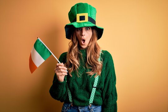 Beautiful patriotic woman wearing green hat holding irish flag celebrating saint patricks day scared in shock with a surprise face, afraid and excited with fear expression