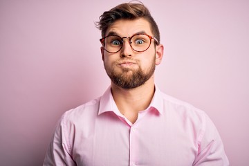Fototapeta premium Young handsome blond man with beard and blue eyes wearing pink shirt and glasses puffing cheeks with funny face. Mouth inflated with air, crazy expression.