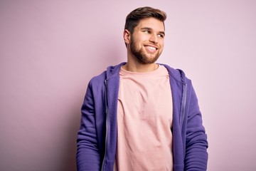Young blond man with beard and blue eyes wearing purple sweatshirt over pink background looking away to side with smile on face, natural expression. Laughing confident.