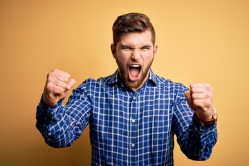 Young blond businessman with beard and blue eyes wearing shirt over yellow background angry and mad raising fists frustrated and furious while shouting with anger. Rage and aggressive concept.