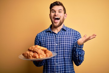 Young blond man with beard and blue eyes holding plate with french croissants to breakfast very happy and excited, winner expression celebrating victory screaming with big smile and raised hands