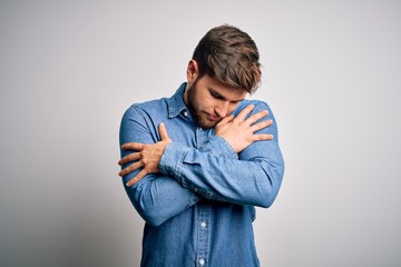 Young handsome blond man with beard and blue eyes wearing casual denim shirt Hugging oneself happy...