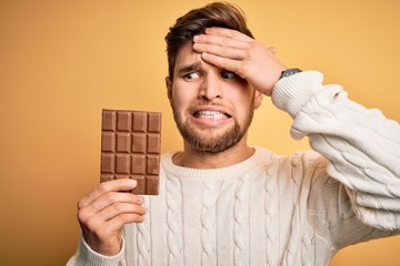 Young blond man with beard and blue eyes holding chocolate bar over yellow background stressed with...