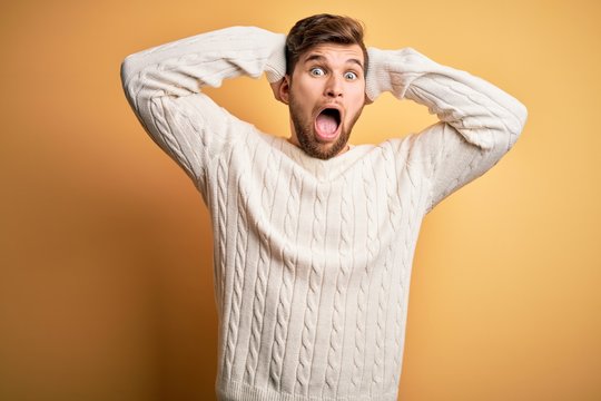 Young blond man with beard and blue eyes wearing white sweater over yellow background Crazy and scared with hands on head, afraid and surprised of shock with open mouth