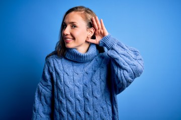 Young beautiful blonde woman wearing casual turtleneck sweater over blue background smiling with hand over ear listening an hearing to rumor or gossip. Deafness concept.