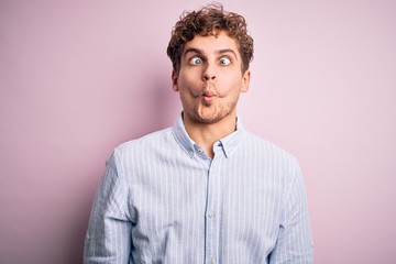 Young blond handsome man with curly hair wearing striped shirt over white background making fish face with lips, crazy and comical gesture. Funny expression.