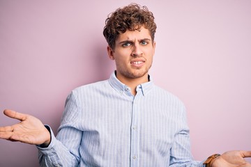 Young blond handsome man with curly hair wearing striped shirt over white background clueless and confused with open arms, no idea concept.