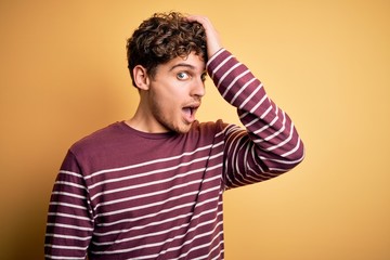 Young blond handsome man with curly hair wearing casual striped sweater surprised with hand on head...
