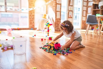 Beautiful toddler wearing glasses and unicorn diadem playing with tractor and building blocks at kindergarten