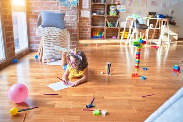Obraz premium Beautiful toddler wearing glasses and unicorn diadem lying down on the floor drawing using paper and pencil at kindergarten