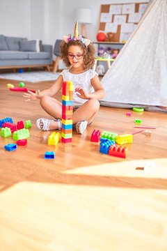 Beautiful toddler wearing glasses and unicorn diadem sitting playing with building blocks smiling at kindergarten