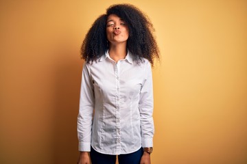 Young beautiful african american elegant woman with afro hair standing over yellow background making fish face with lips, crazy and comical gesture. Funny expression.