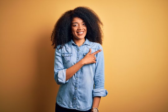Young beautiful african american woman with afro hair standing over yellow isolated background cheerful with a smile of face pointing with hand and finger up to the side happy and natural