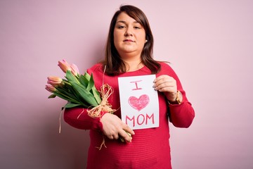 Beautiful plus size woman celebrating mothers day holding message and bouquet of tulips with a confident expression on smart face thinking serious