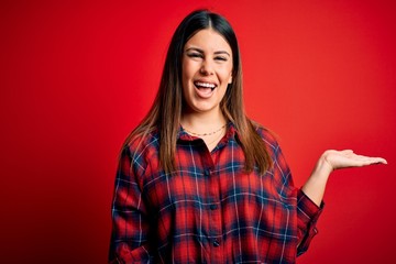 Fototapeta premium Young beautiful woman wearing casual shirt over red background smiling cheerful presenting and pointing with palm of hand looking at the camera.