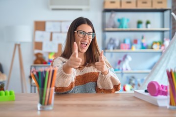 Young beautiful teacher woman wearing sweater and glasses sitting on desk at kindergarten pointing fingers to camera with happy and funny face. Good energy and vibes.