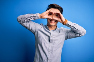 Young handsome chinese man wearing casual shirt standing over isolated blue background Doing heart shape with hand and fingers smiling looking through sign